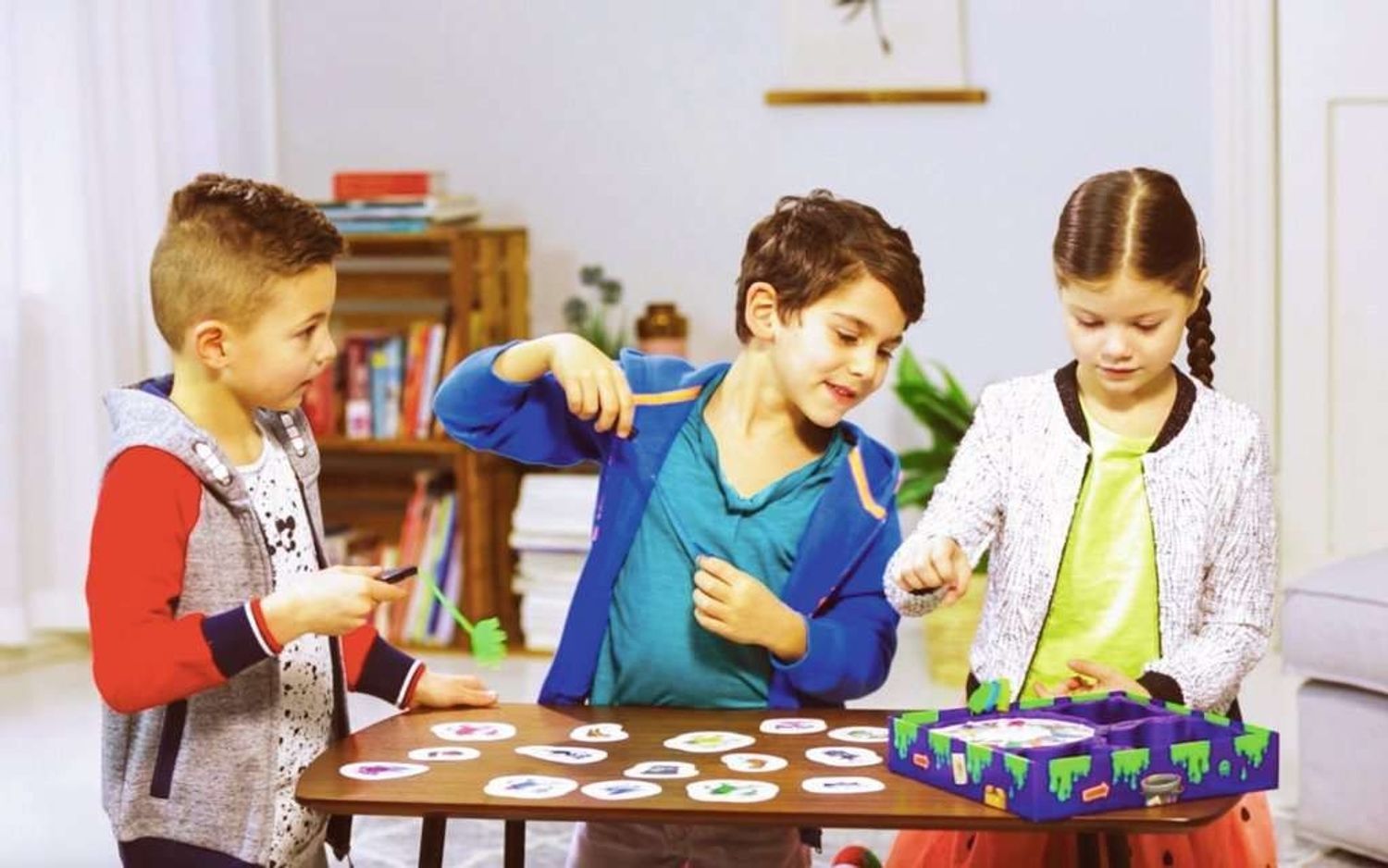 Drie kinderen spelen. Eén kind houdt een grijpgereedschap vast. Bord en kaarten op tafel. Monsterillustraties.