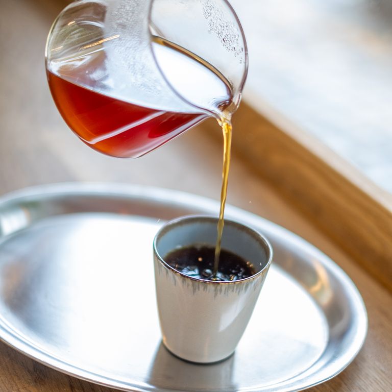 Du café est versé dans une tasse. Soucoupe argentée. Plateau de table en bois clair.