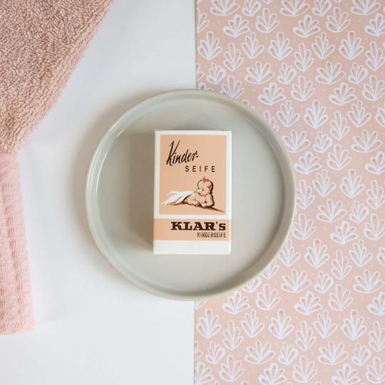 Klar-Seifen Kinderseife verpakking op een bord. Roze achtergrond met patroon. Handdoek links.