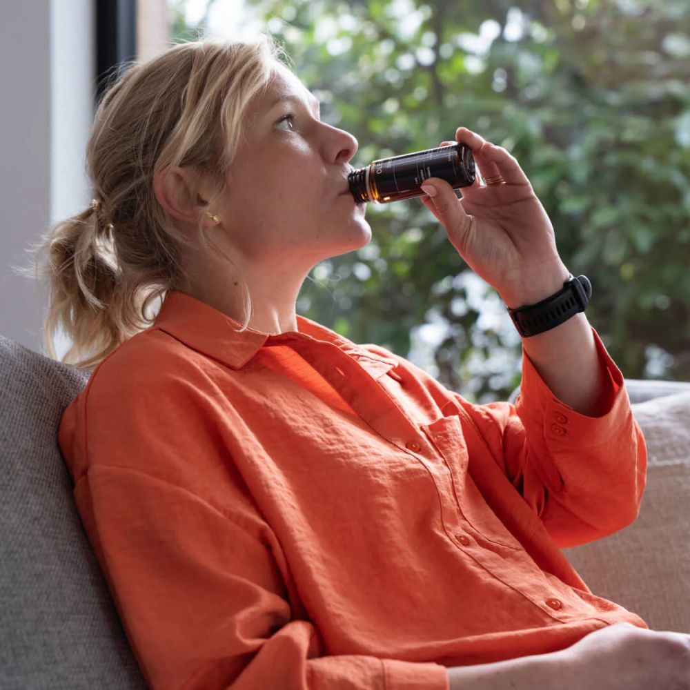 Vrouw drinkt uit een fles. Opschrift: OLEUS. Interieur met raam op de achtergrond.