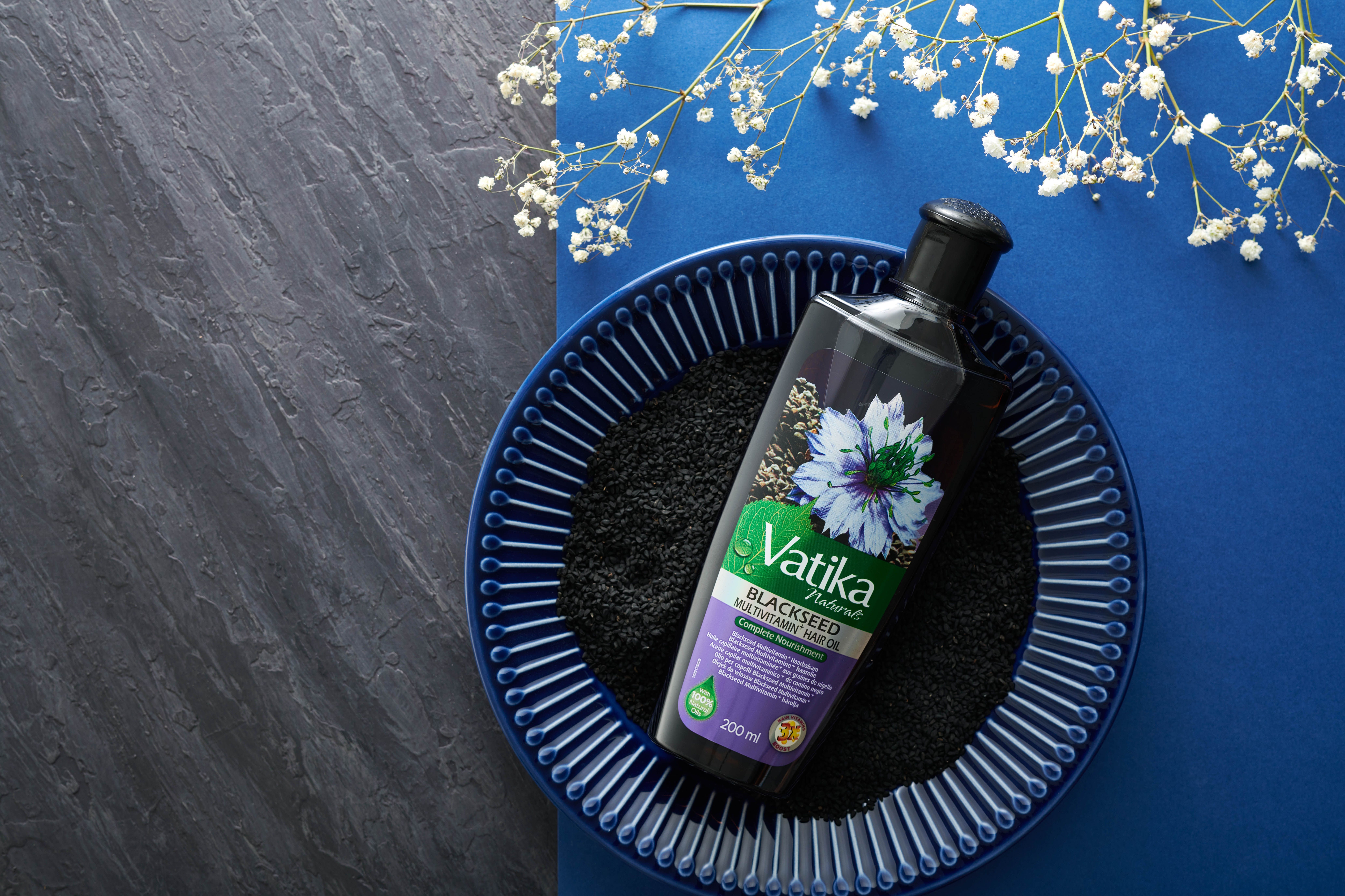 Vatika Naturals Schwarzkümmel haarolie op blauw bord met zwarte komijn. Op blauwe en grijze achtergrond.