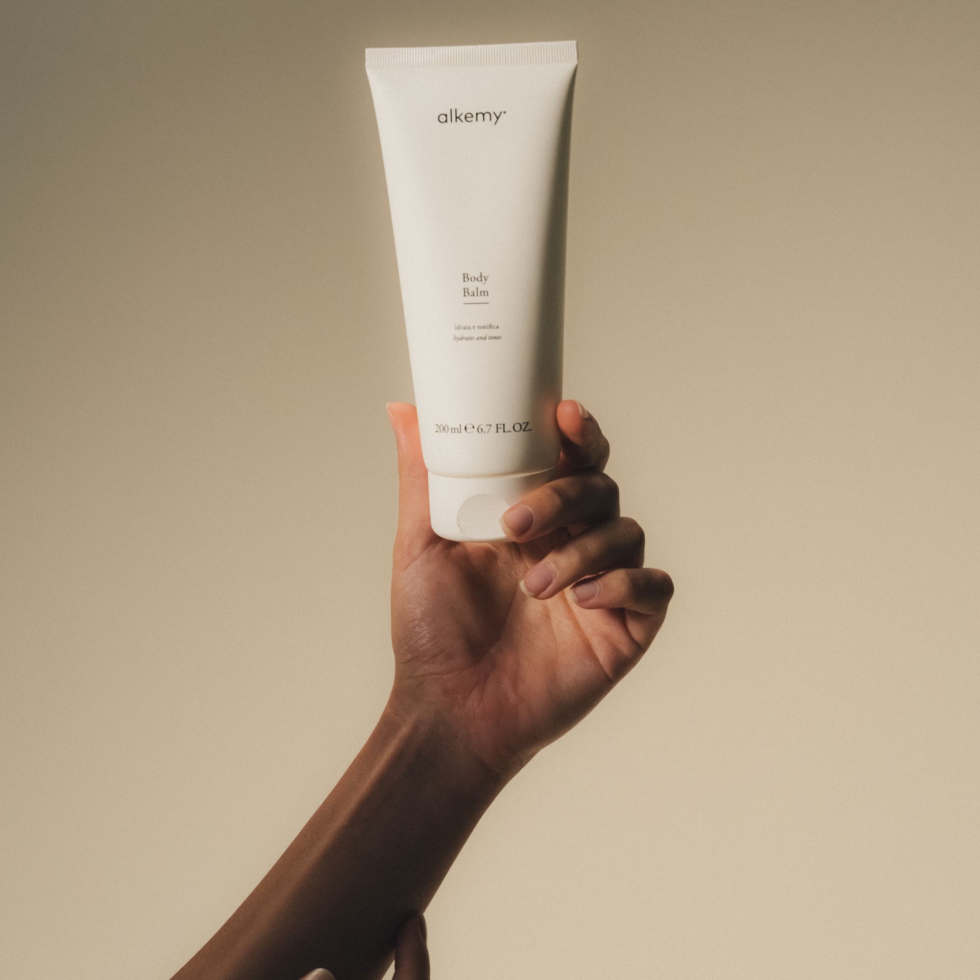 Une main tient un tube blanc de baume corporel. Le tube a un bouchon rabattable et le logo 'alkemy'.