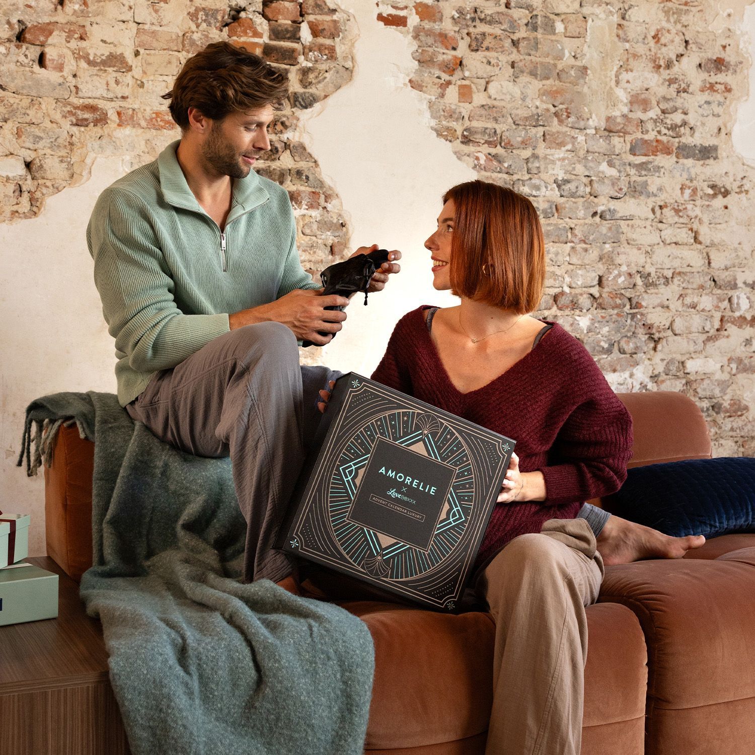 Een man en een vrouw zitten samen. De vrouw houdt een adventskalenderbox vast. AMORELIE-logo zichtbaar.