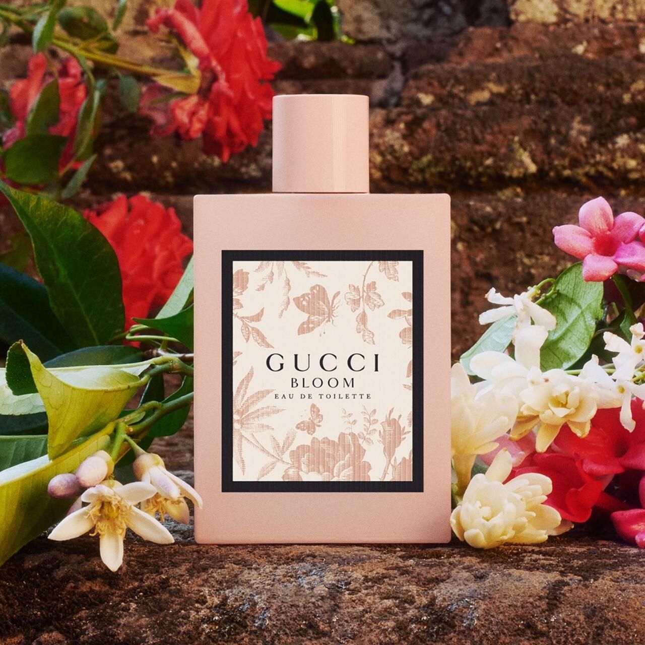 Roze fles met bloemenprint, voor stenen muur en bloemen. Opschrift: Gucci Bloom Eau de Toilette.