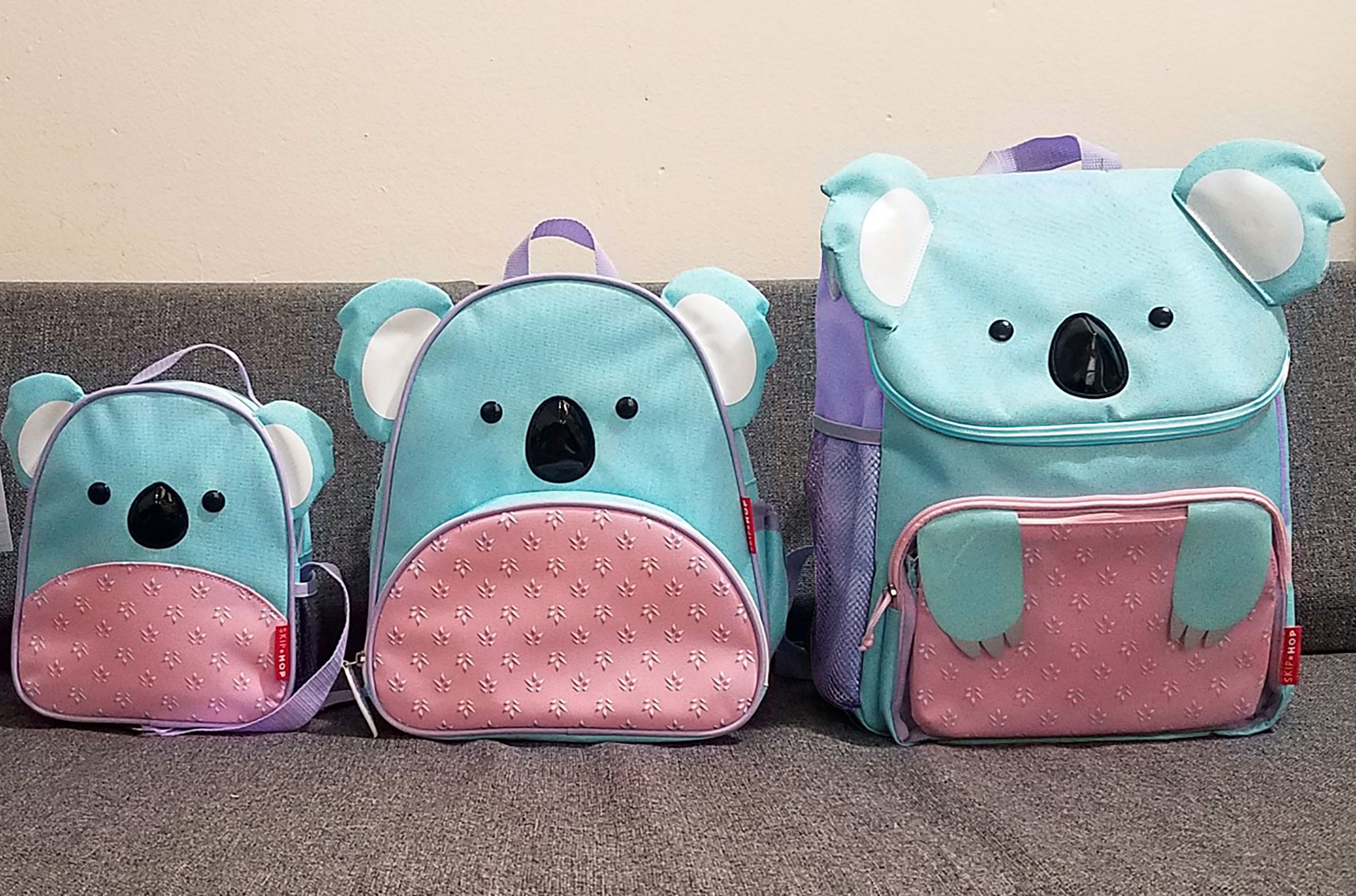 Drie Skip Hop Zoo Koala rugzakken. Verschillende maten. Blauw en roze. Op grijze bank.