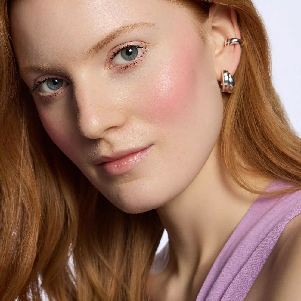 Portrait d'une femme avec du blush rose sur les joues. Elle a les cheveux roux et porte des boucles d'oreilles.