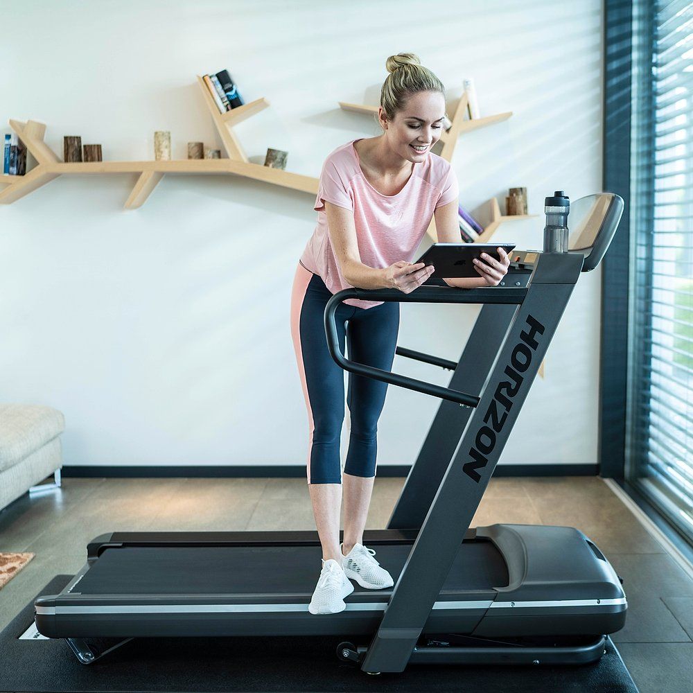 Femme debout près d'un tapis roulant tenant une tablette. Tapis roulant avec logo Horizon. Vêtements de sport.