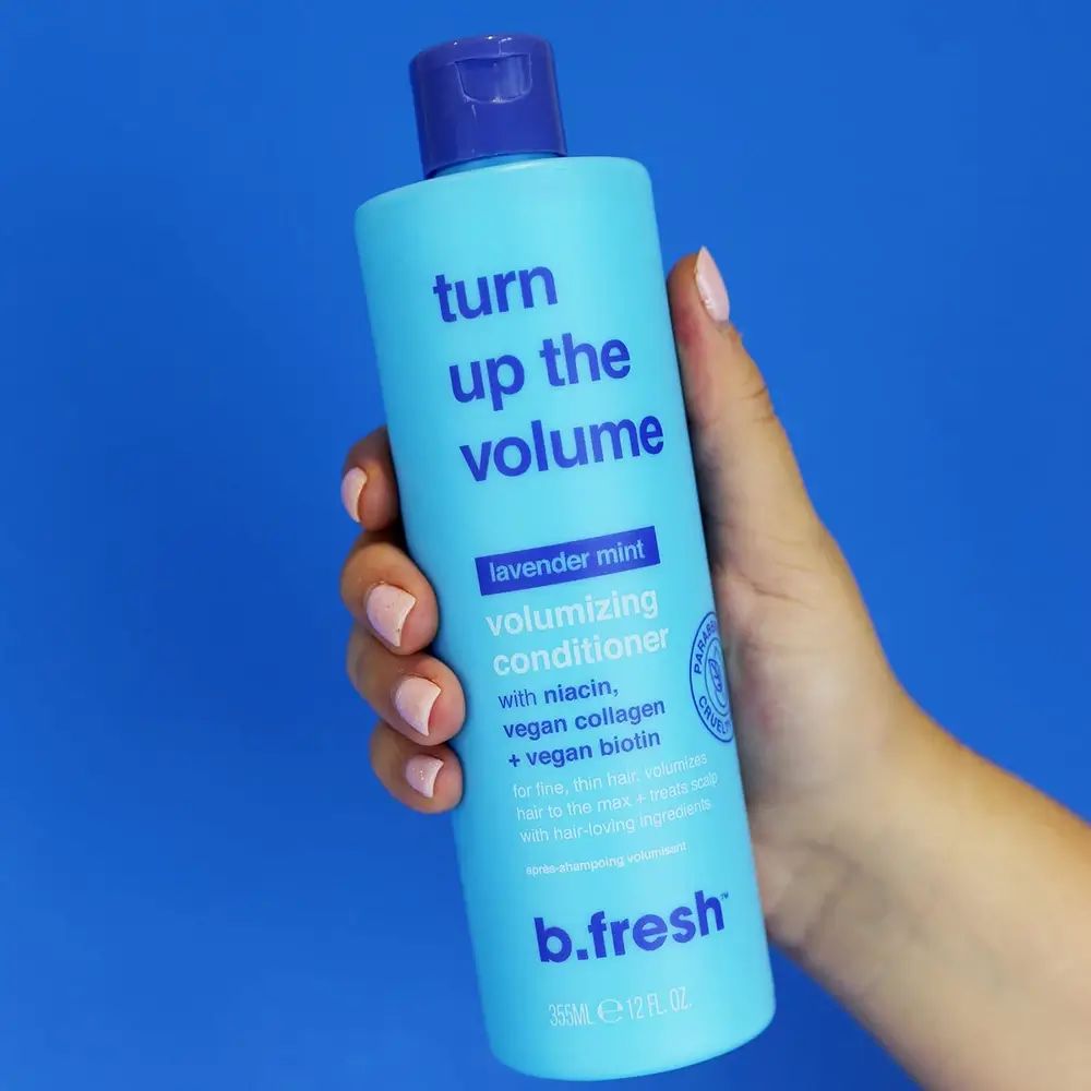 Lichtblauwe fles met blauwe dop, vastgehouden door een hand. Opschrift: Turn Up the Volume, Volumizing Conditioner. Bevat niacine, vegan collageen, biotine.