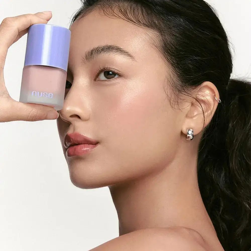 Een vrouw houdt een flesje vloeibare blush voor haar gezicht. De fles is roze en heeft een paarse dop. De vrouw heeft donker haar en draagt oorbellen.