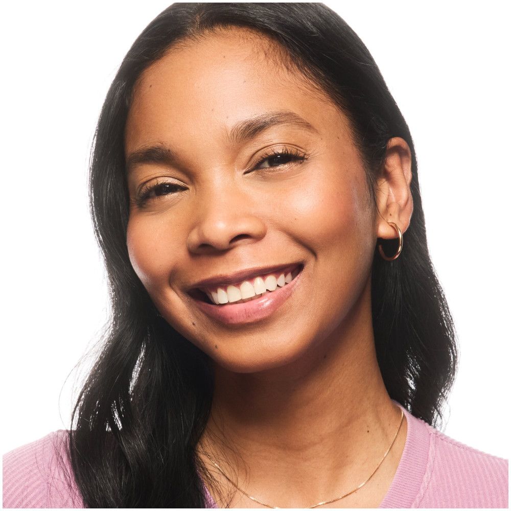 Portret van een lachende vrouw. Ze draagt foundation en heeft een egale teint. Gouden oorringen.