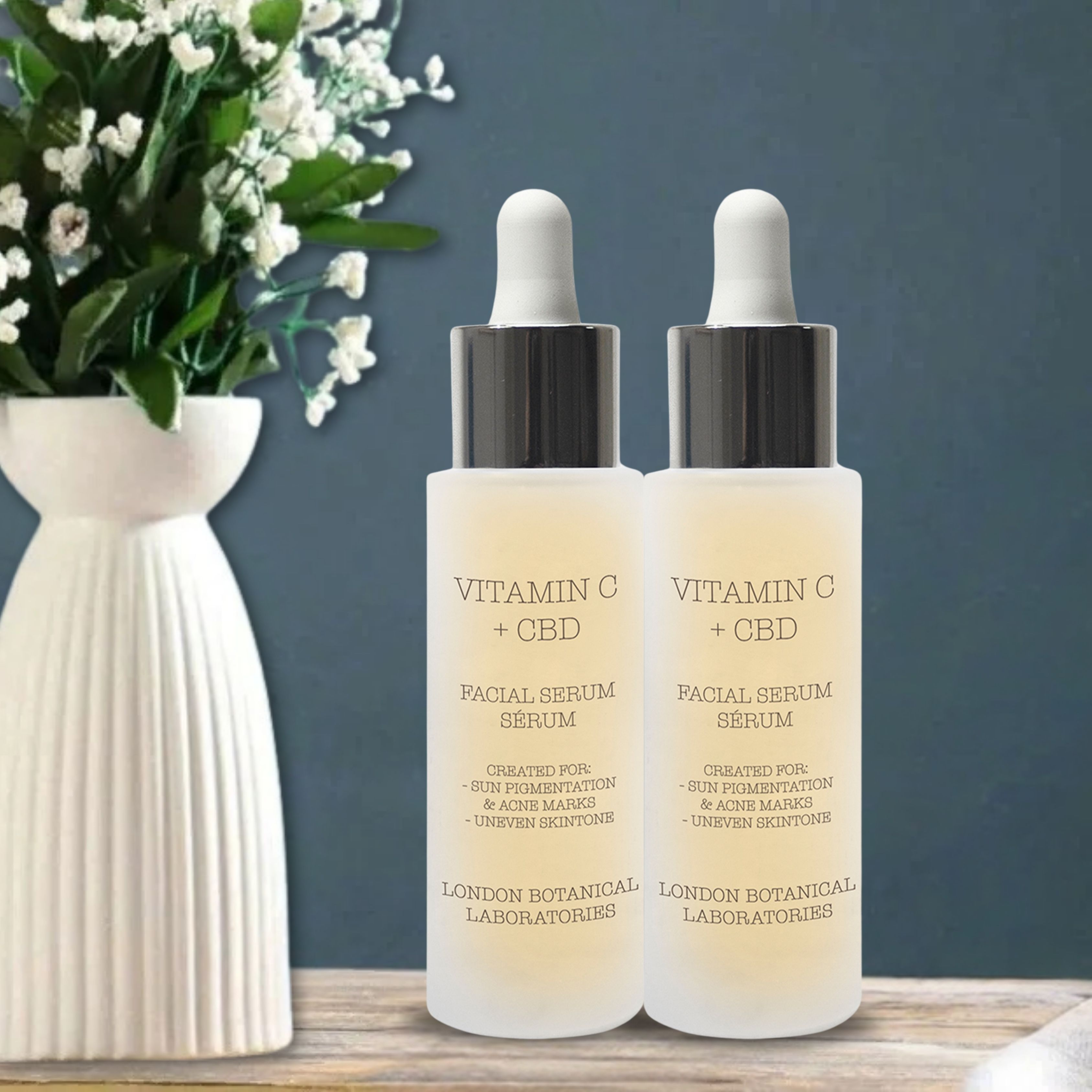 Twee serumflessen met pipetten. Opschrift: Vitamin C + CBD. Merk: London Botanical Laboratories. Achtergrond: vaas met bloemen.