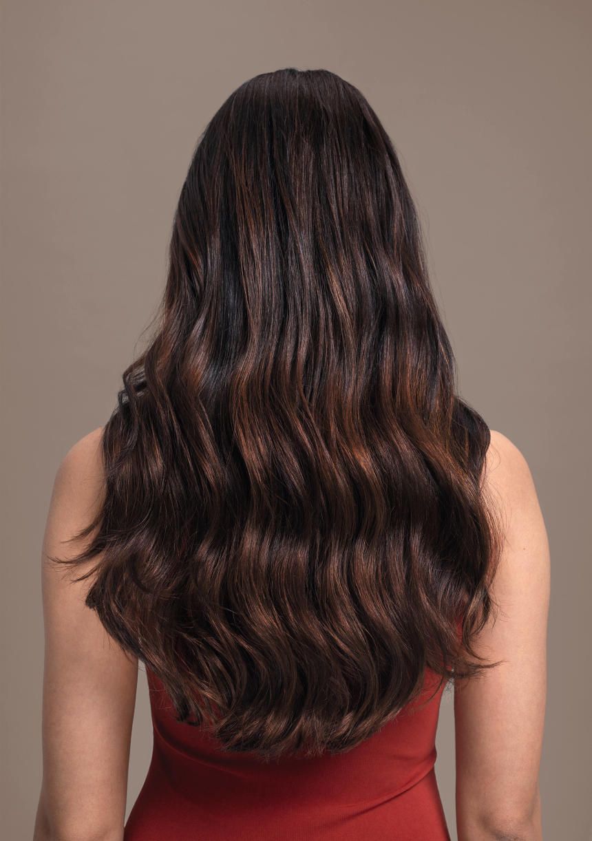 Vue arrière d'une femme aux cheveux bruns ondulés. Les cheveux sont mi-longs et présentent des reflets.