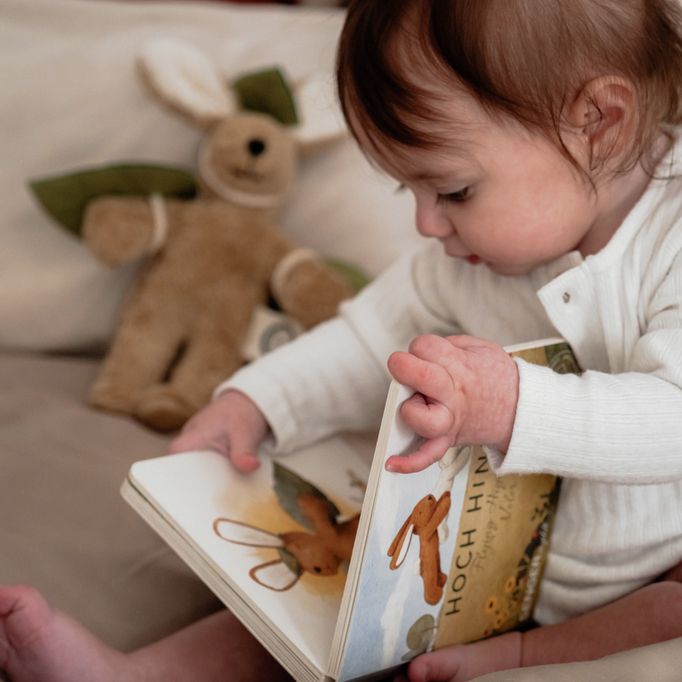 Bébé lisant un livre. Livre avec illustrations de lapins. Titre : Hoch Hinaus. Peluche lapin en arrière-plan.