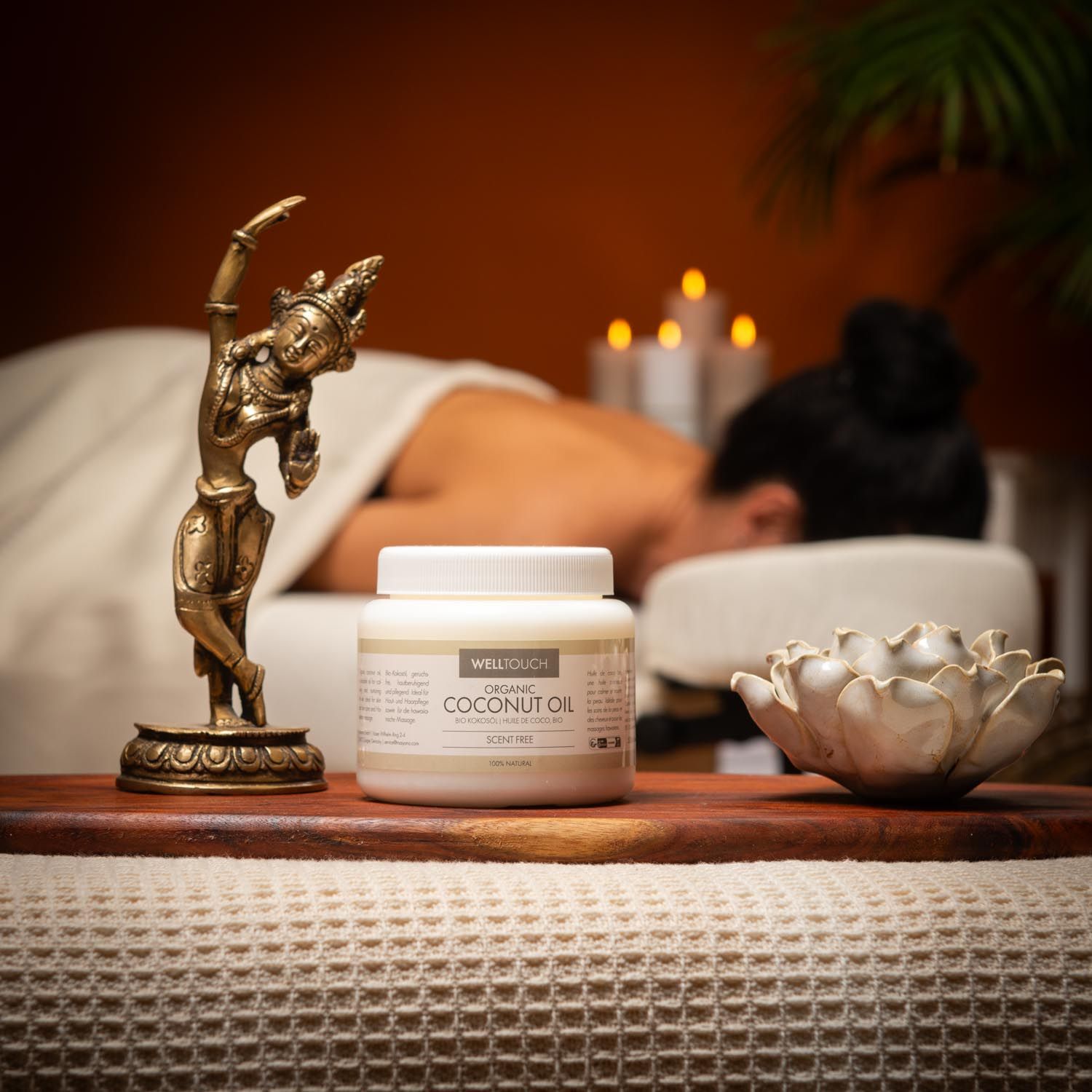 Wit glazen pot met COCONUT OIL. Massage in uitvoering. Bronzen beeld en lotusbloem op houten plank.