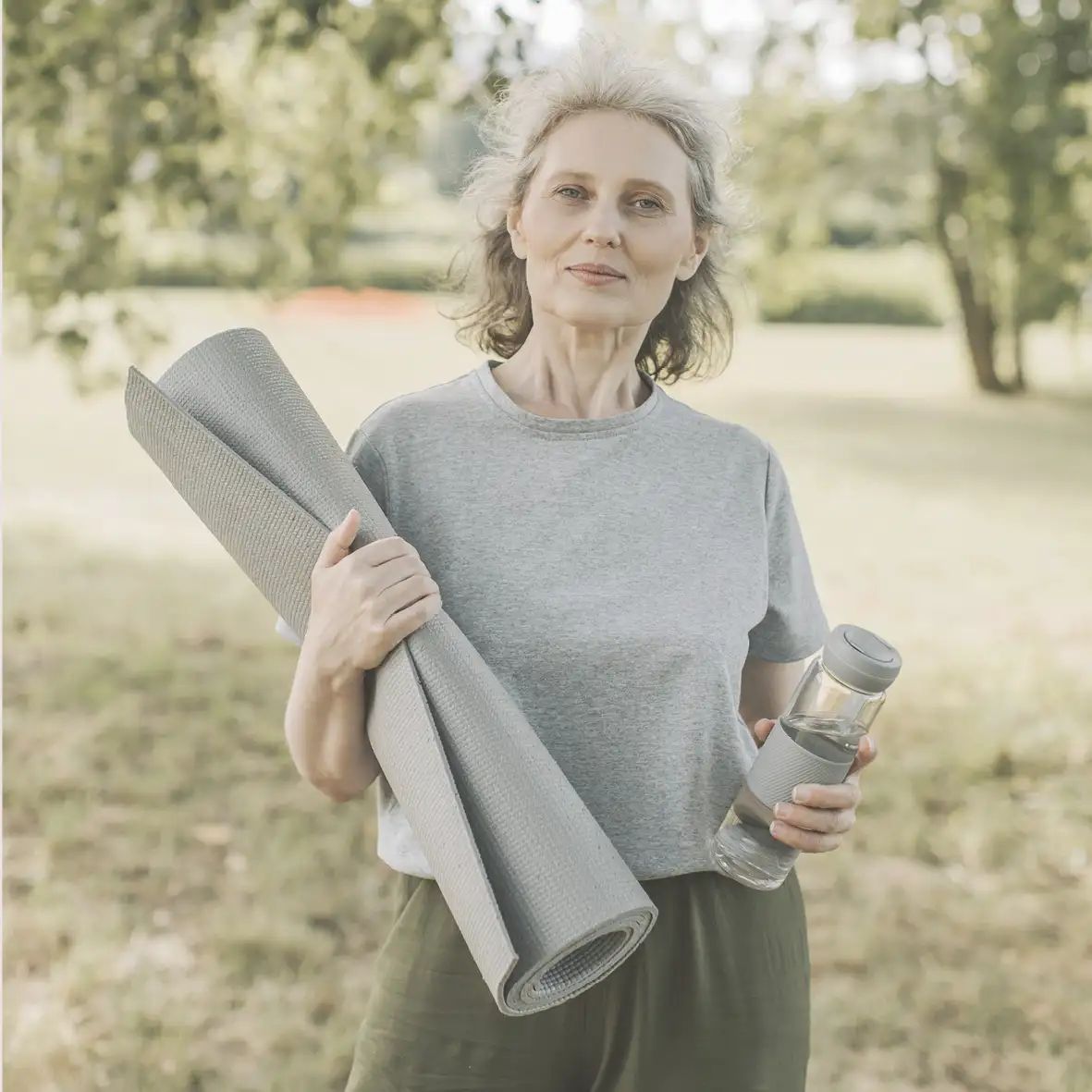 Vrouw met yogamat en waterfles in het park. Ze draagt een grijs T-shirt en een groene broek.