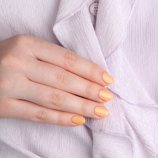 Main avec ongles vernis couleur pêche pastel. Ongles courts et vernis. Fond : matière claire et texturée.