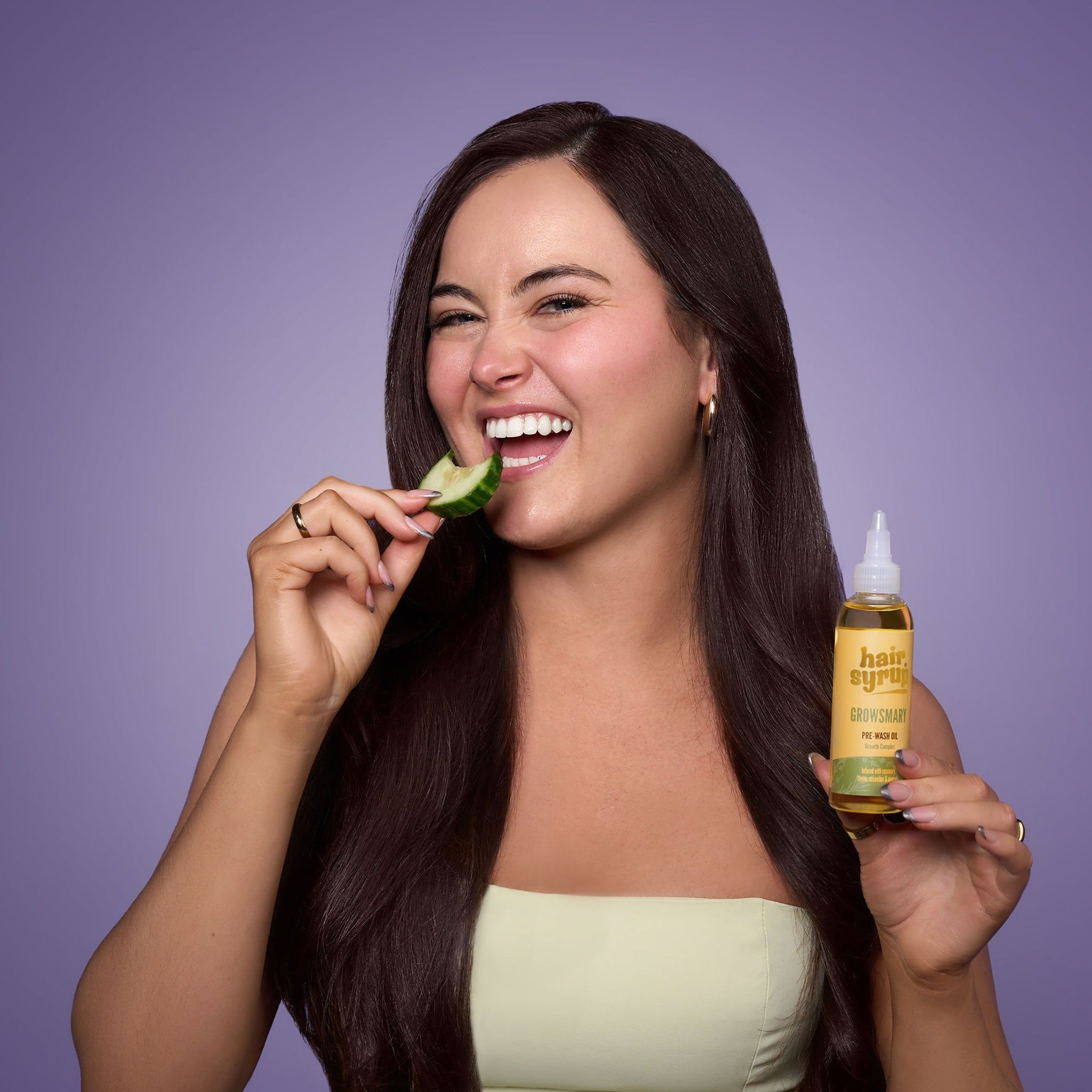 Femme aux longs cheveux foncés tenant une bouteille « Hair Syrup Growsmary » et mangeant un concombre. Sur fond violet.