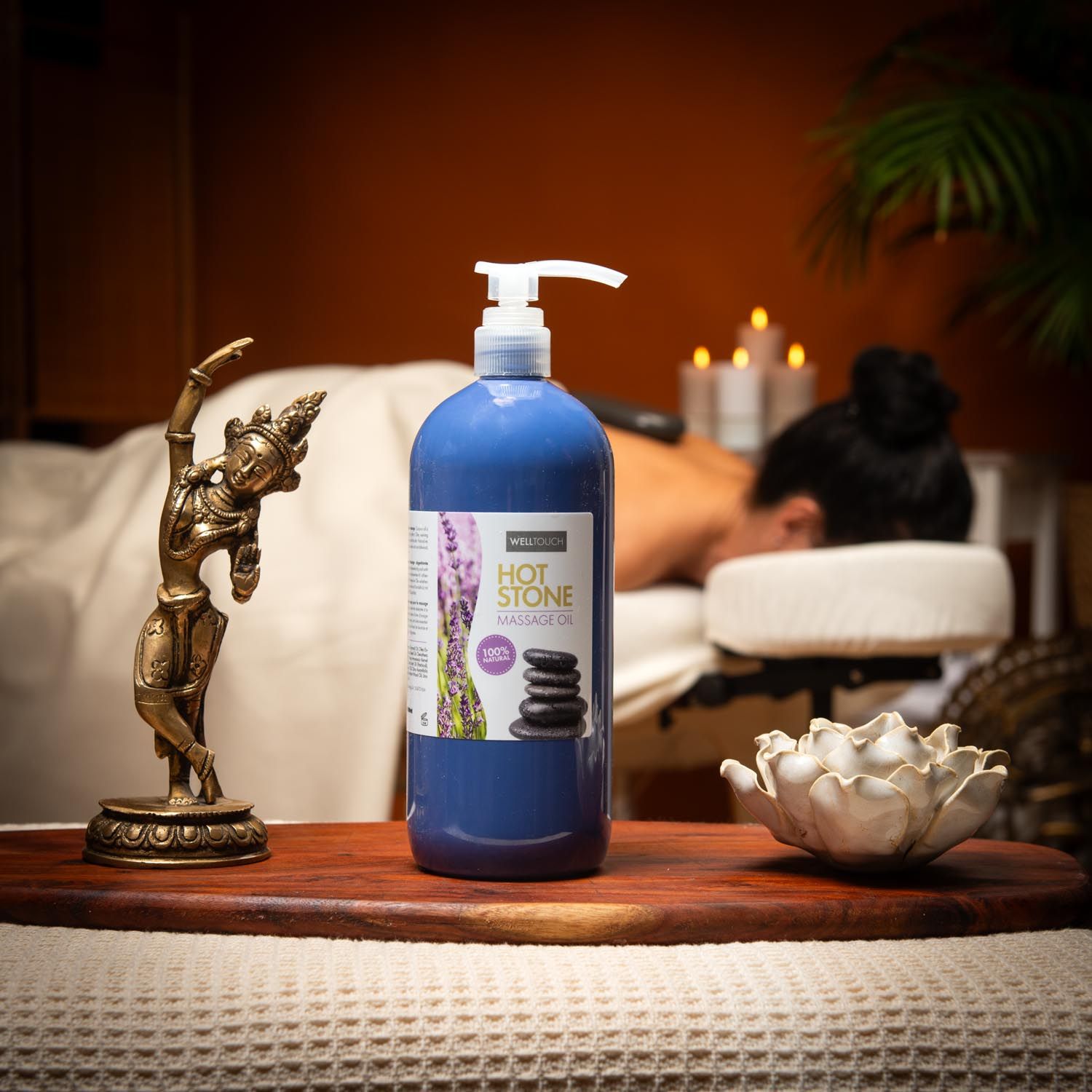 Blauwe fles Welltouch Hot Stone Massageöl met pomp. Op houten dienblad, naast Boeddha-beeld en lotusbloem. Achtergrond: persoon op massagetafel.