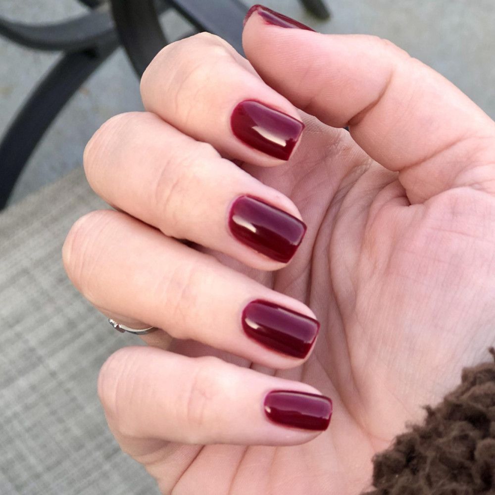 Main avec ongles vernis rouge foncé. Forme carrée, finition brillante. Bague au doigt.