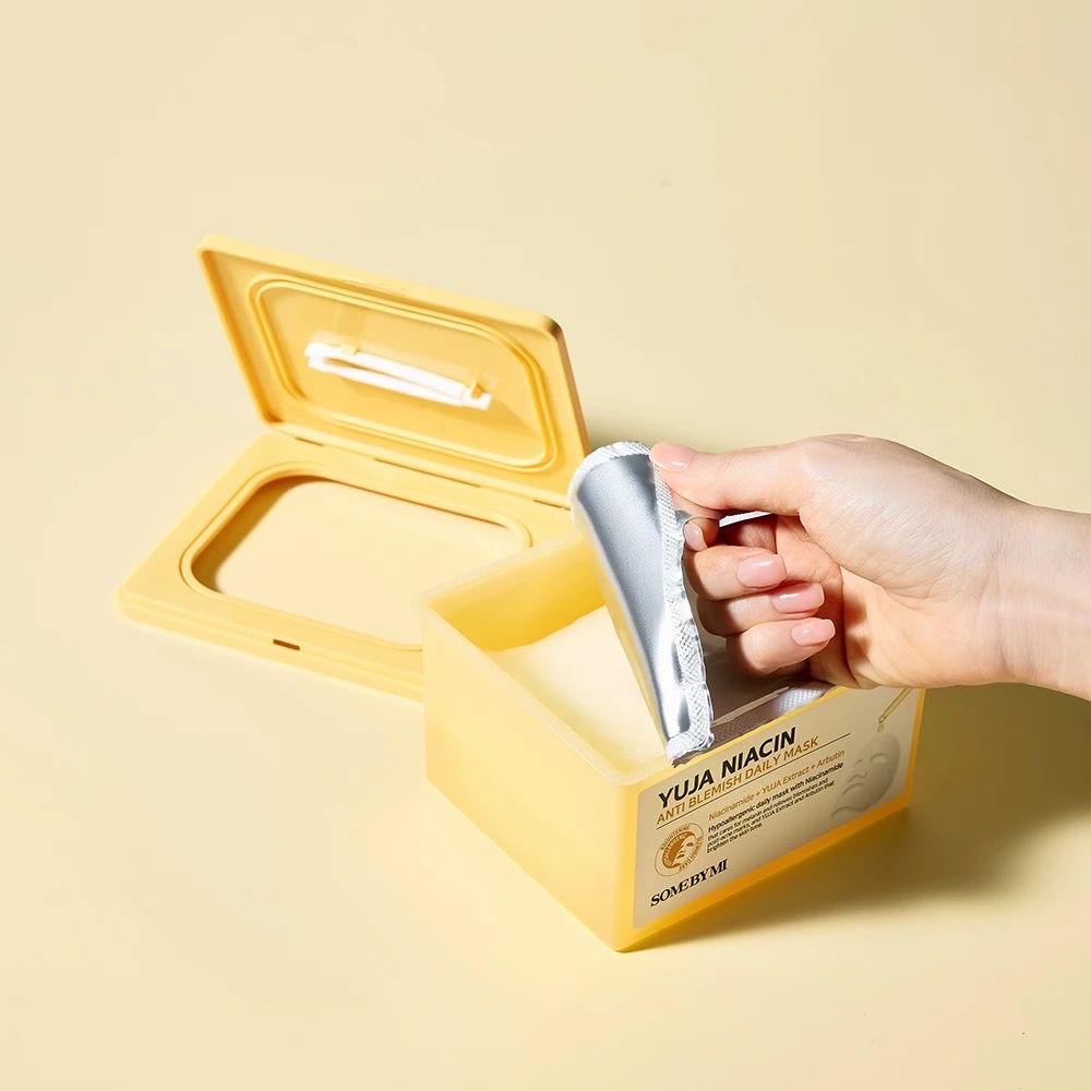 Open gele maskerdoos. Een hand trekt een zilveren masker verpakking eruit. Opschrift: Yuja Niacin.