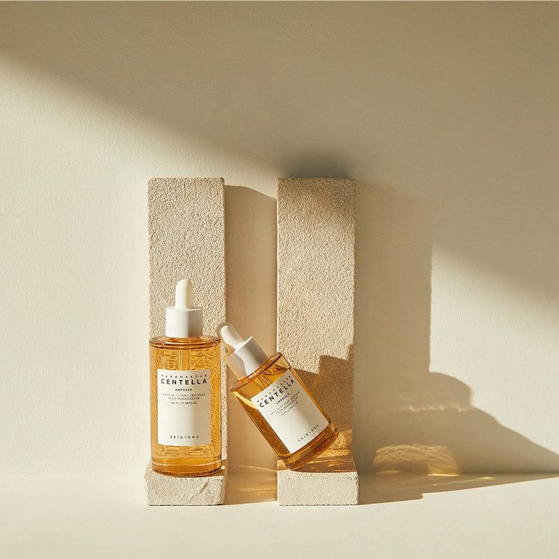 Twee flessen met geelachtige vloeistof en witte dop. Opschrift: MADAGASCAR CENTELLA AMPULE. SKIN1004. Op sokkels.