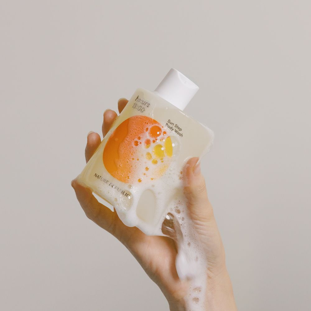 Een hand houdt een fles douchegel vast. Schuim stroomt uit de fles. Tekst en een oranje ontwerp zijn zichtbaar op de fles.