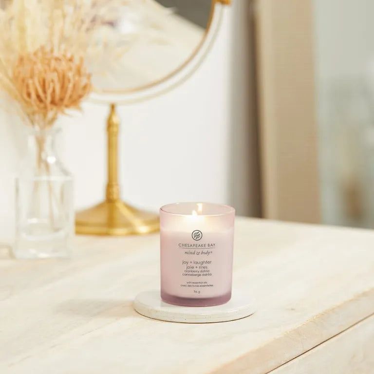 Bougie rose Chesapeake Bay mind & body, joy + laughter, cranberry dahlia, sur un dessous de plat. Miroir et vase en arrière-plan.