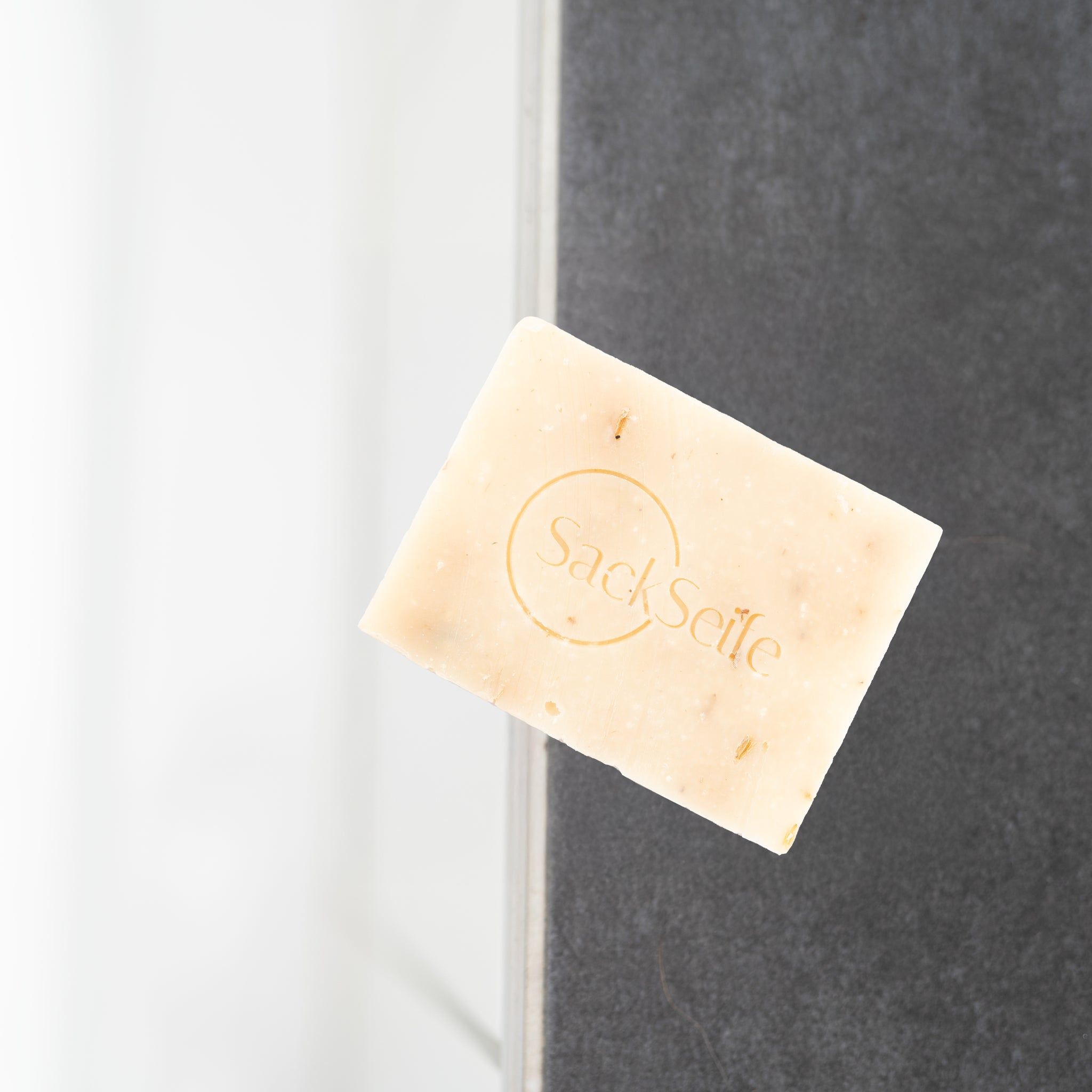 Savon rectangulaire avec logo SackSeife. Couleur claire, particules visibles. Posé sur un mur gris.