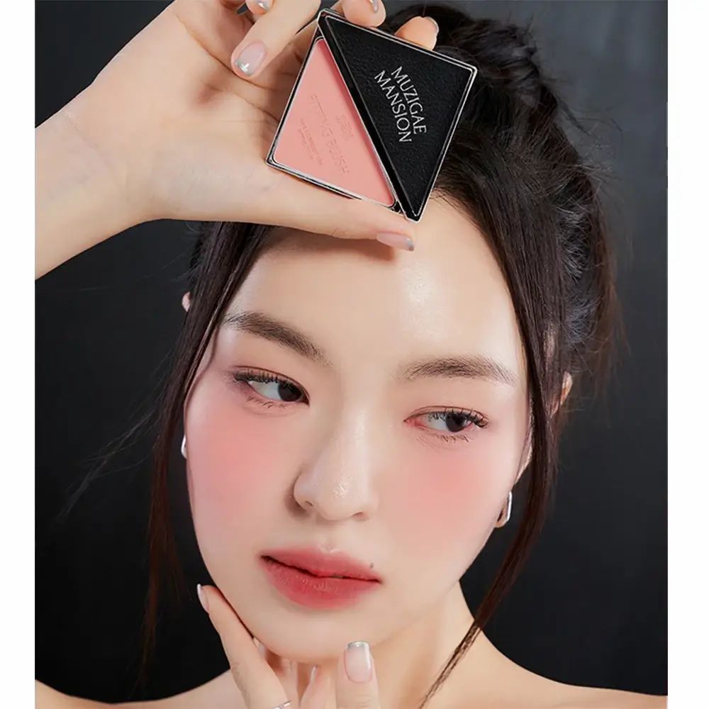 Vrouw houdt blush-palette vast. Zwarte palette met MUZIGAE MANSION-logo. Roze blush op de wangen van de vrouw.