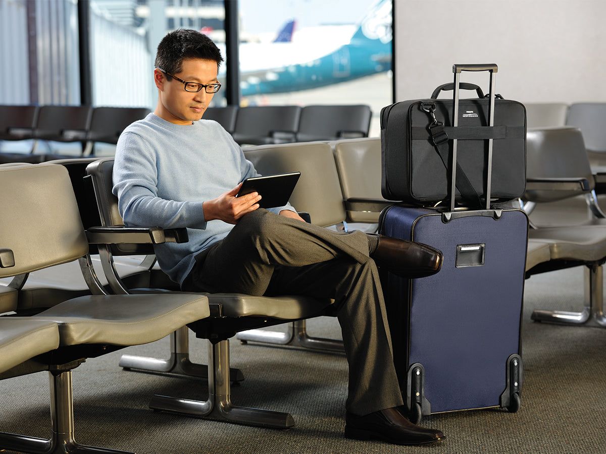 Homme assis dans une salle d'attente d'aéroport avec un sac de voyage noir sur une valise à roulettes à côté de lui.