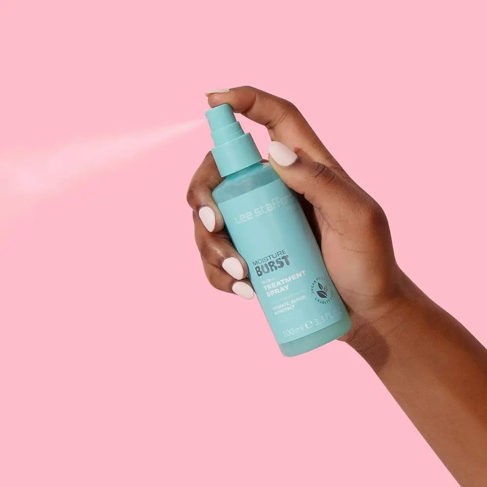 Hand houdt turquoise sprayfles vast. Opschrift: Lee Stafford, Moisture Burst, 10-in-1 Treatment Spray. Spuitnevel zichtbaar.