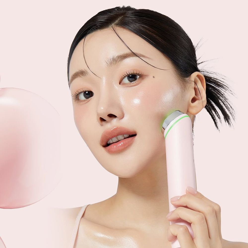 Femme utilisant un appareil de soin de la peau. Boîtier rose, lumière verte. Nom du produit non visible.