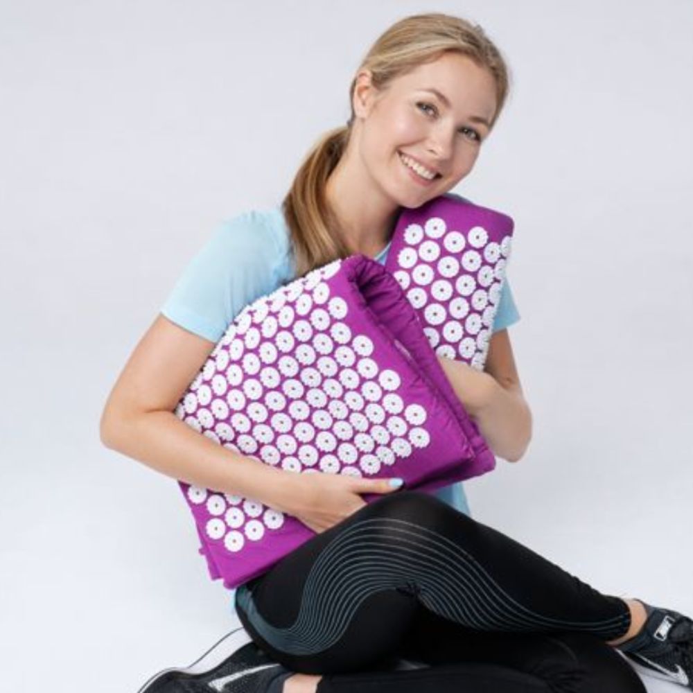 Femme tenant un kit d'acupression. Tapis et coussin visibles. Elle sourit.