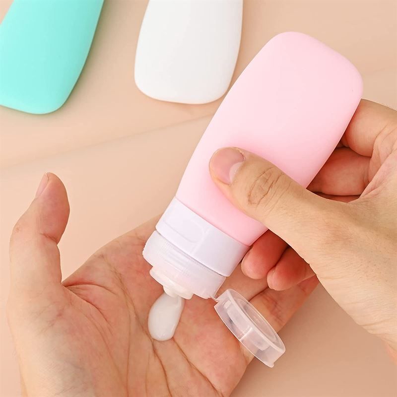 Roze reisflacon met witte dop. Een hand houdt de fles vast, waaruit een vloeistof komt. Dop ernaast.