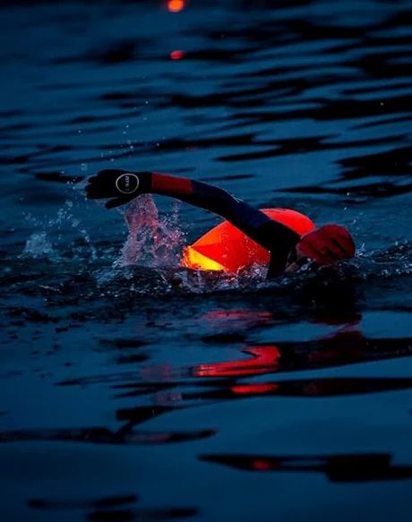 Personne nageant dans l'eau avec une bouée orange. Environnement sombre, l'eau reflète la lumière.