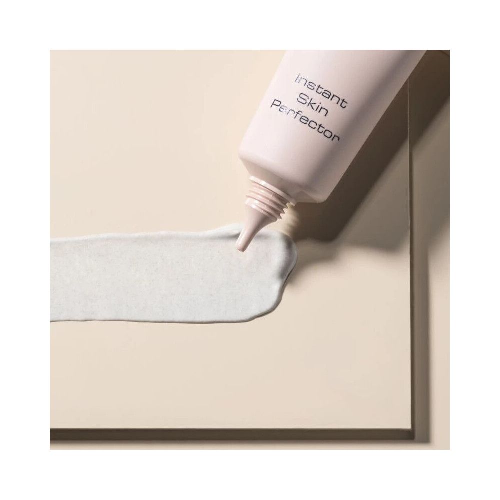 Tube de produit et goutte. Inscription Instant Skin Perfector. Tube rose, pointe. Goutte vert clair. Fond beige.