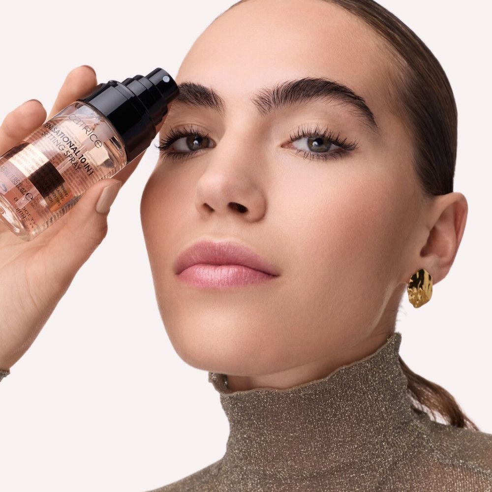 Vrouw houdt setting spray voor gezicht. Productnaam en kenmerken zichtbaar. Gouden kraag en oorbellen.