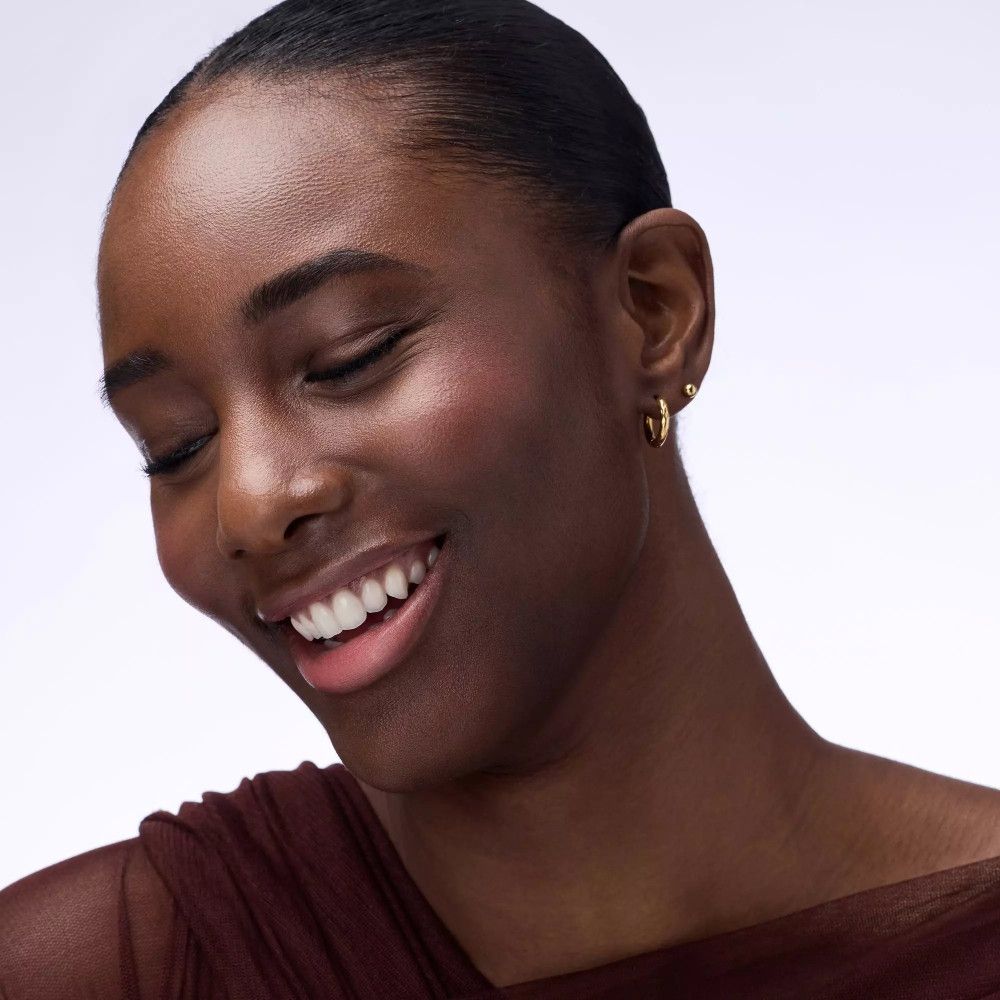 Portrait d'une femme souriante avec du blush. Elle porte des créoles dorées. Le teint est bronzé et naturel.