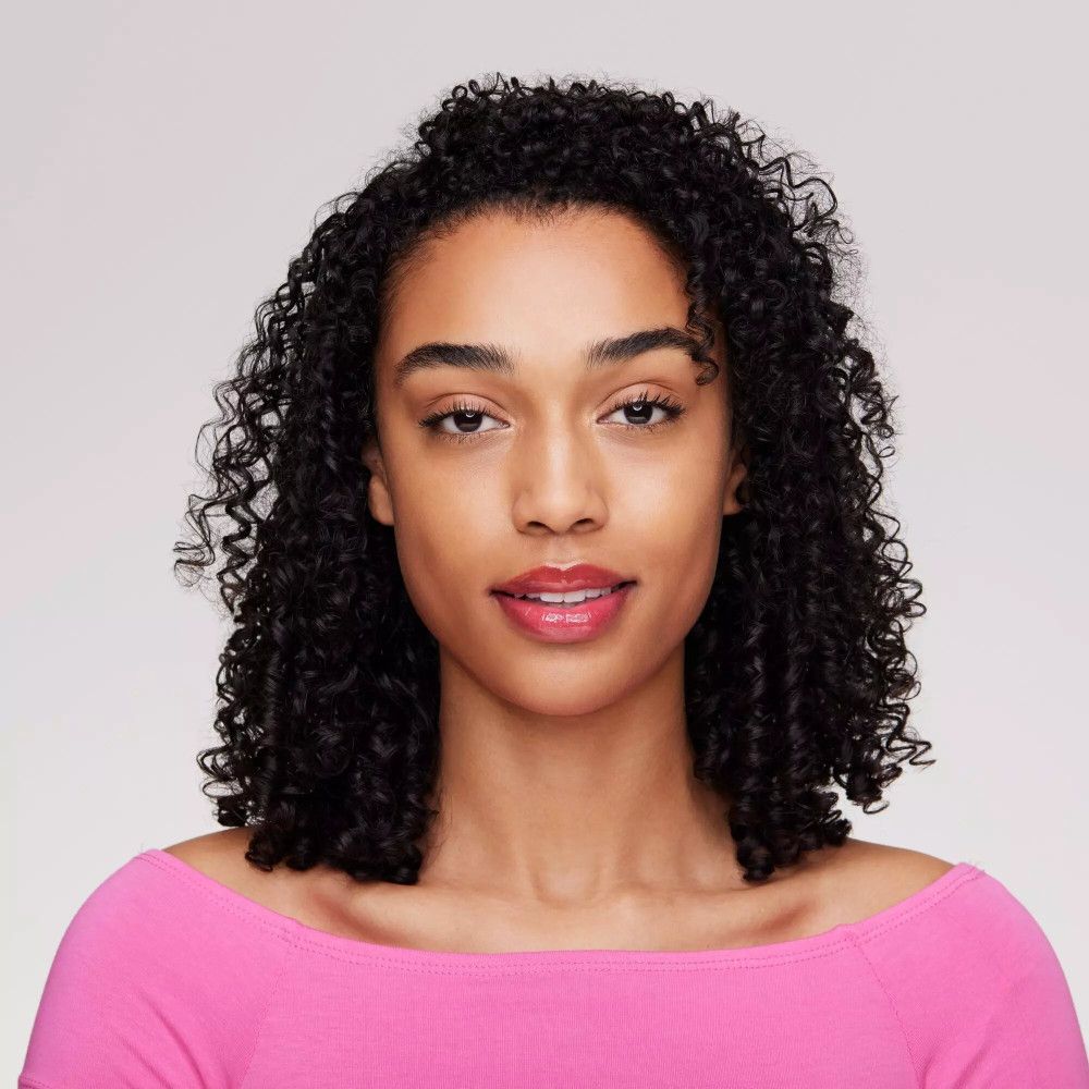 Portrait d'une femme aux cheveux bouclés. Elle porte du rouge à lèvres rose. Le fond est gris clair.