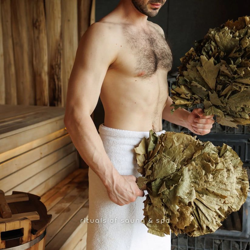 Man houdt twee bossen bladeren vast. Sauna-elementen op de achtergrond.