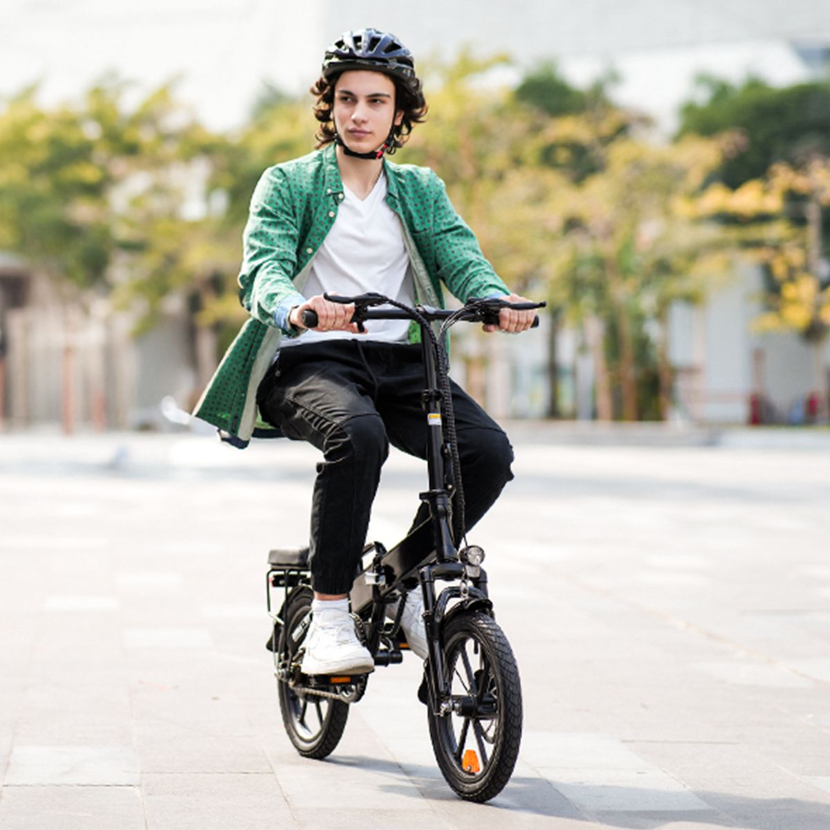 Persoon fietst op een zwarte, opvouwbare elektrische fiets. Draagt helm en groene jas. Achtergrond: bomen.