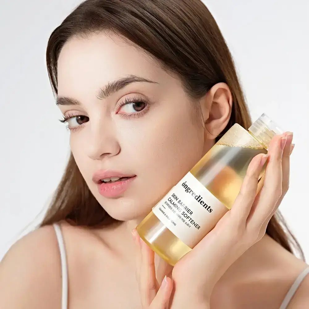 Vrouw houdt een fles ONGREDIENTS Skin Barrier Calming Softener vast. Productlabel duidelijk zichtbaar. Vrouw kijkt in de camera.