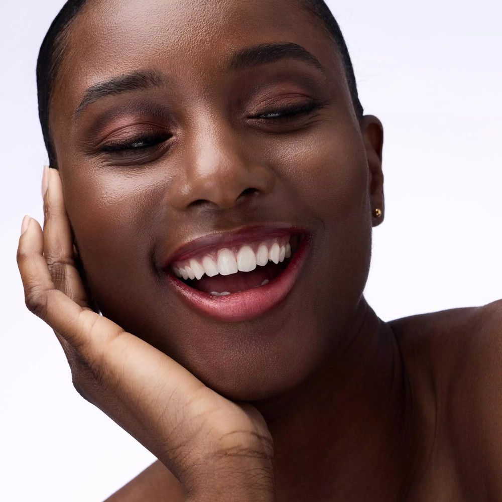 Portrait d'une femme souriante avec fard à paupières dans les tons marron et orange. Elle a la peau foncée et tient son menton.