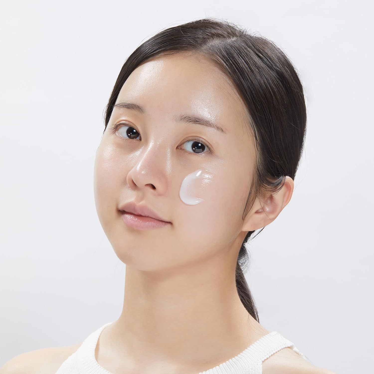 Gros plan d'une femme appliquant de la crème sur son visage. Peau claire, cheveux foncés. Débardeur blanc.