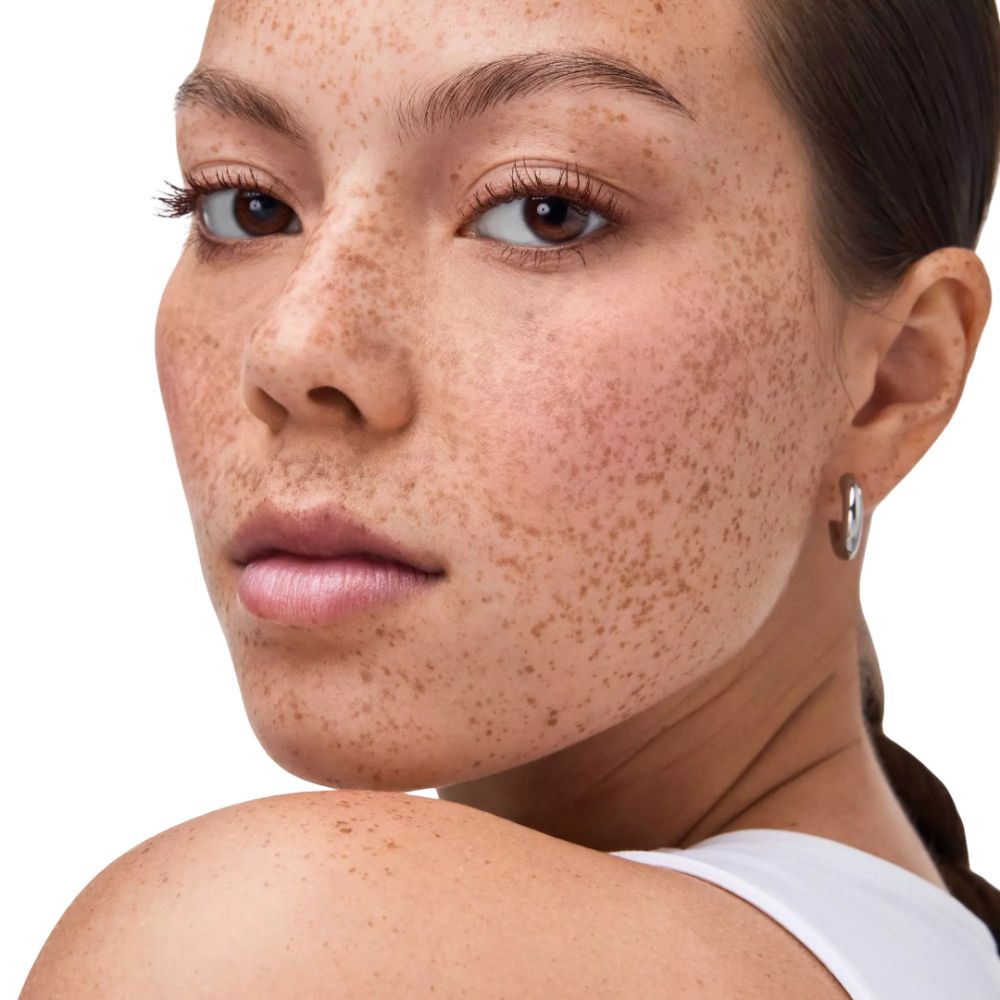 Gros plan d'une femme avec des taches de rousseur. Elle porte un débardeur blanc et des boucles d'oreilles. Regard vers la caméra.