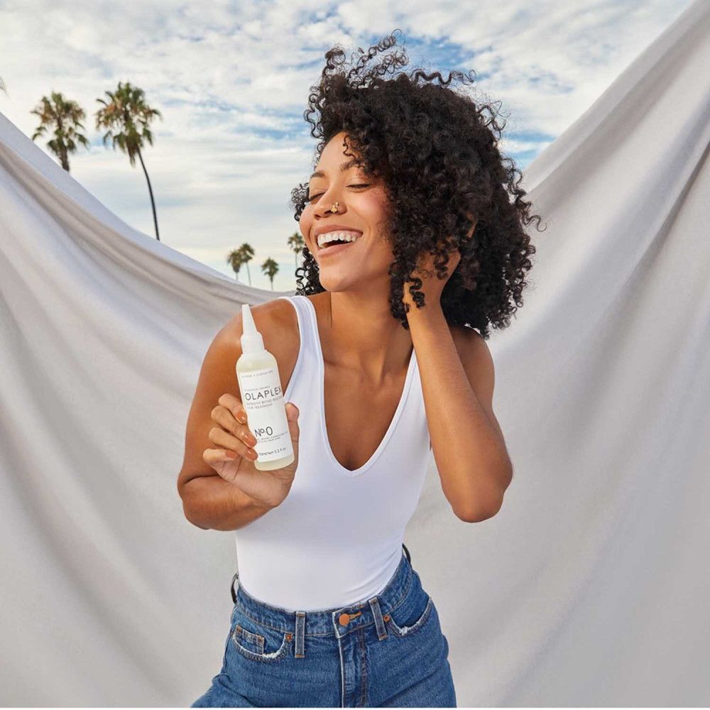 Femme tenant un flacon OLAPLEX No. 0. Inscription : OLAPLEX Intensive Bond Building Hair Treatment. Fond : plage.