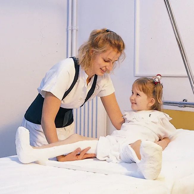 Kind met been in het gips, ondersteund door een vrouw met ruggordel. Ligt op een bed.