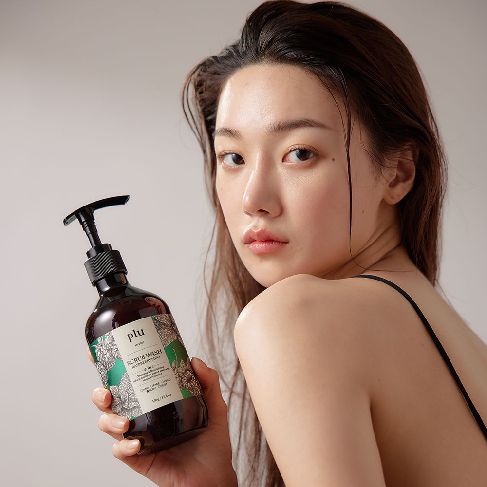 Une femme tient une bouteille marron avec une pompe noire. Inscription : PLU Scrub Wash Raspberry Mint.