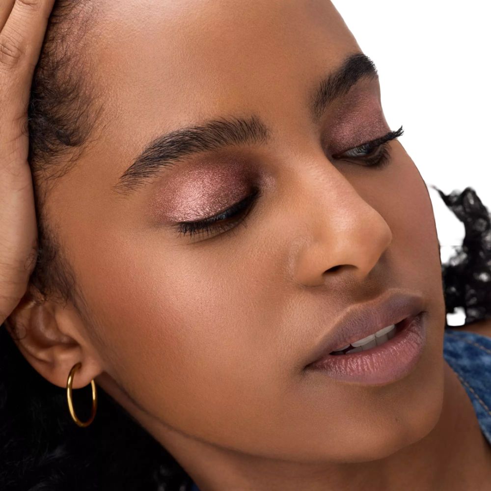 Portrait d'une femme avec fard à paupières rose. Créoles dorées. Elle pose sa tête sur sa main. Fond blanc.