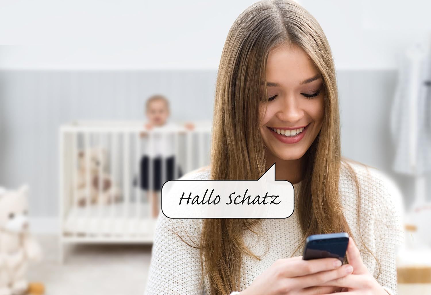 Vrouw met smartphone, tekstballon met tekst 'Hallo schatz'. Babybedje op de achtergrond, babyfoon niet zichtbaar.